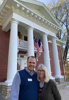 New owners Jack and Monica standing by red brick house with white pillars and flags hanging on house.