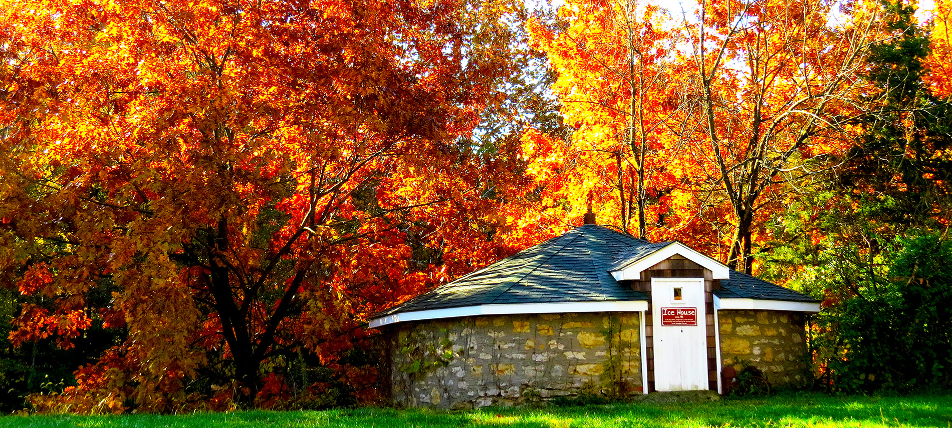 A squat brown stone building with a white door in front of trees with brilliant red and yellow leaves