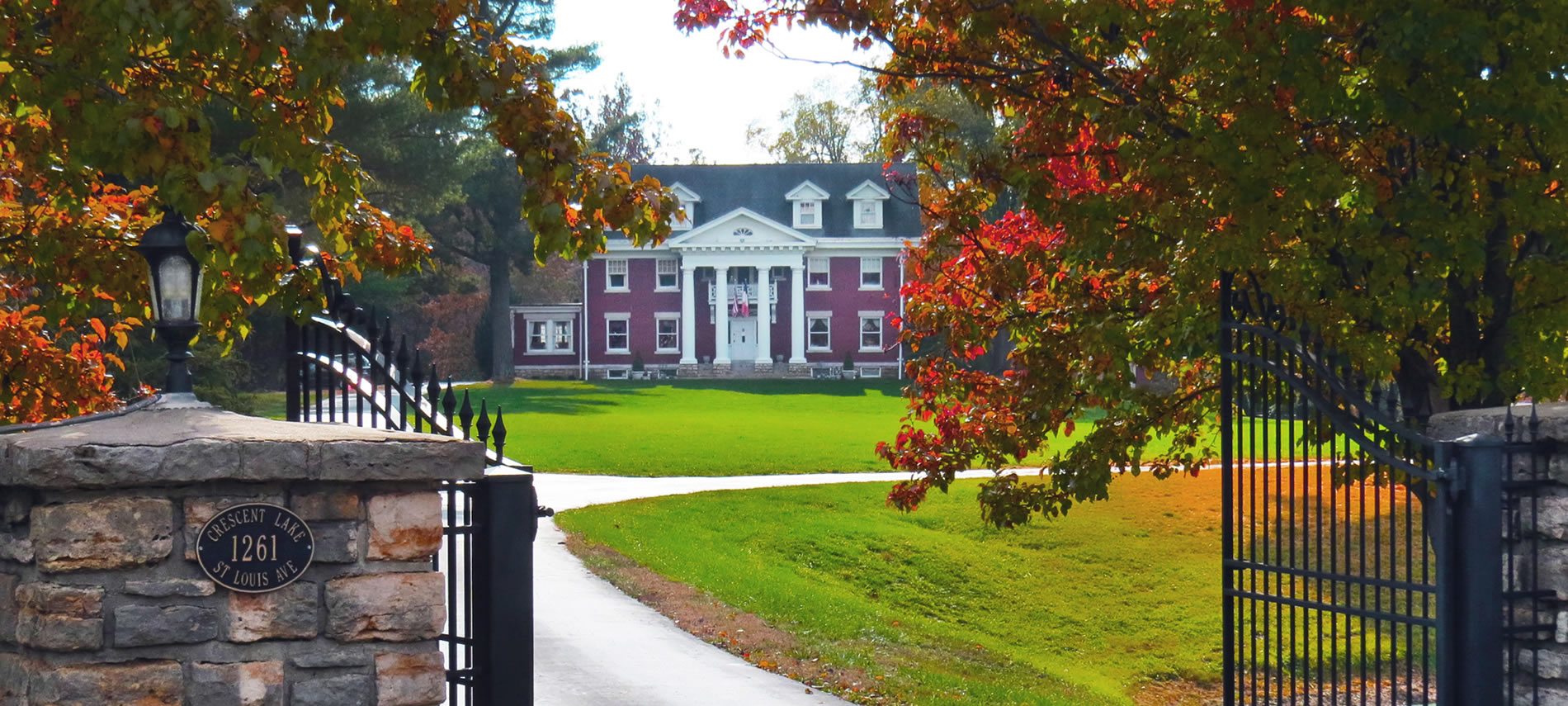 A gated entrance to a driveway with a red brick mansion with four white columns visible in the distance