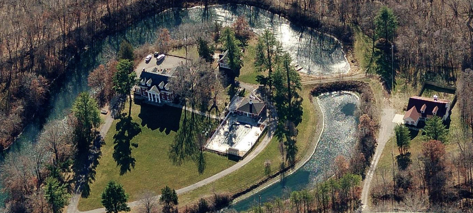 Aerial view with water surrounding a red brick mansion and pool, with a white house in a clearing to the right